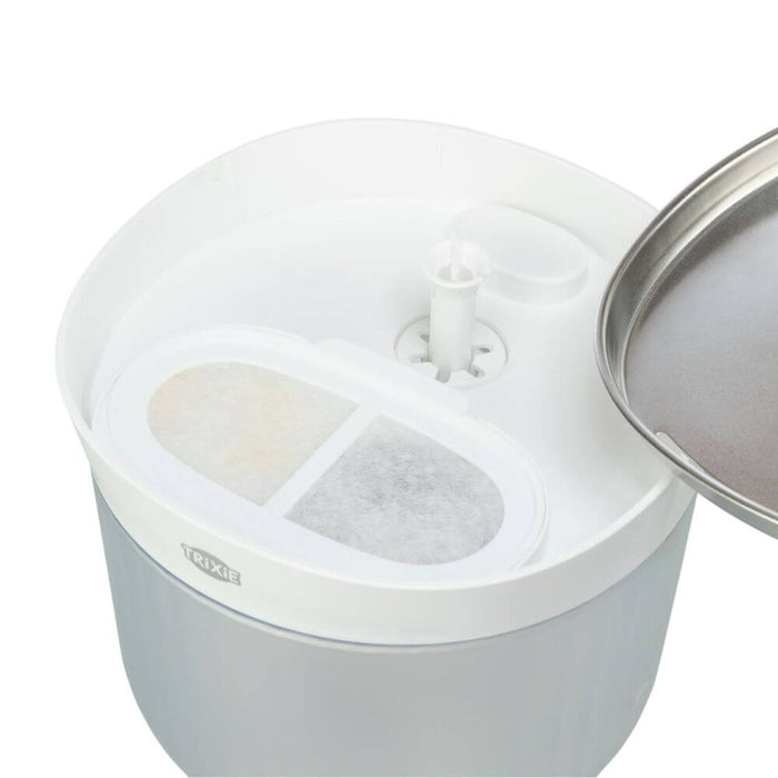 A close-up of a white and light gray pet waterer or feeder container with a stainless steel lid partially visible on the right. The top has a central white spout and two oval filter compartments, one showing a beige filter and the other a gray filter. It is a dog water bowl with fountain, stainless steel bowl, filters and clear container.